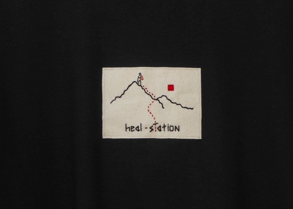 Close-up of a black crewneck t-shirt featuring a rectangular embroidered patch on the chest with a mountain sketch, a red flag, and the text 'heal station'. The neck label shows the brand 'ORCHIN'.